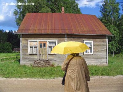 sateenvarjoa pitelevä nainen ränsistyneen hirsitalon edessä auringonpaisteessa