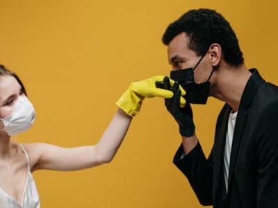 mies hengityssuojain päässään, suojahanska kädessään, suutelee maskiin pukeutuneen naisen kumikäsineeseen verhottua kättä