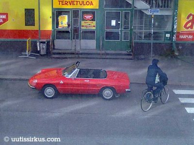 punainen avoauto kadun varressa, vierestä menee huppupäinen pyöräilijä