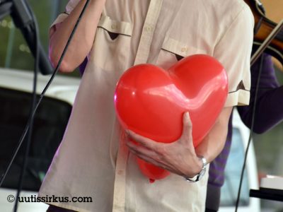 mies mikrofonissa, hänen toisessa kädessään iso sydämenmuotoinen ilmapllo, jota hän painaa rintaansa vasten