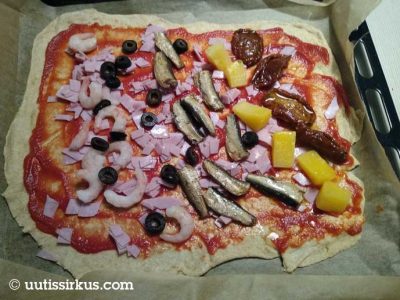 pizza uunipellillä odottaa paistoa - täytteinä kinkkua, katkarapuja, kilohailia, tummia oliiveja ananasta ja aurinkokuivattua tomaattia