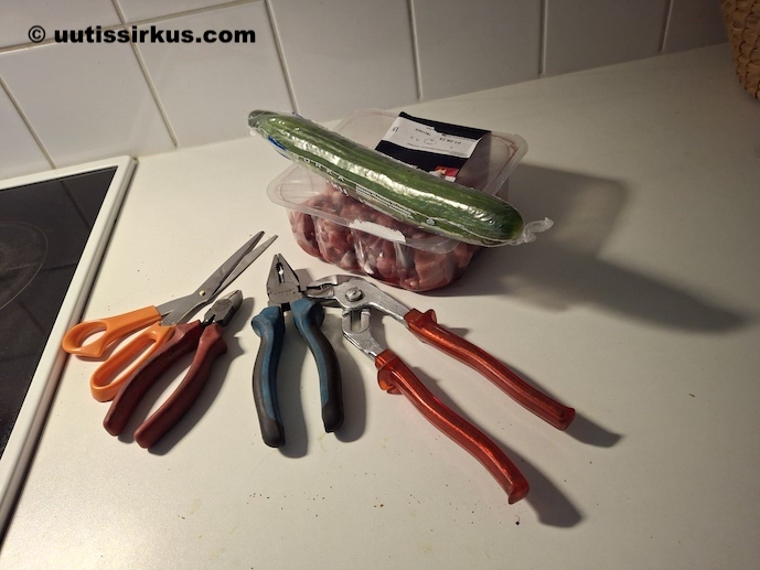 muovinen karjalanpaistilihapakkaust, jonka päällä kurkku muovikääresssään, näiden vieressä pöydällä sakset, kahdenlaiset pihdit ja putkitongit