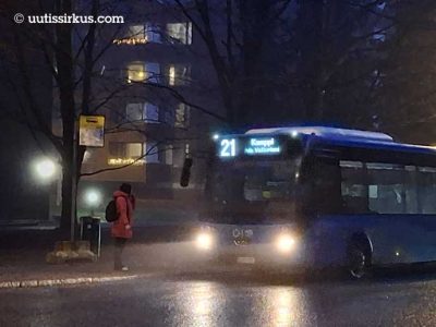 HSL:n sininen bussi numero 21 lähestyy tihkusateisessa sumussa kerrostalon edustalla pysäkkiä, jolla seisoo punatakkinen nainen