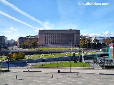 eduskuntatalo ja Kansalaistori Oodin suunnalta nähtynä, takana sininen taivas, taloun takaa tulee kaksi lentokoneen pakokaasuvanaa