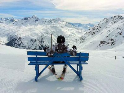 Laskettelija istuu vasrusteissaan rinteen ylimmässä kohdassa sinisellä penkillä selkä kohti kameraa, hänen edessään avautuu näkymä vastapäisille rinteille ja alas osittain pilvien peritossa olevaan laaksoon