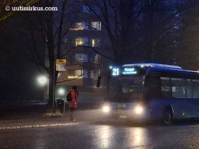 bussi 21 lähestyy pysäkkiä sumuisessaa iltahämärässä, pysäkillä seisoo punaiseen toppatakkiin pukeutunut nainen, taustalla 70-luvun kerrostalo, jonka ikkunoissa valoja