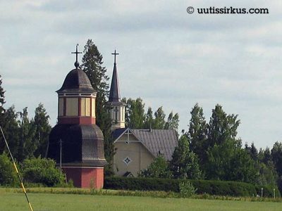 Suomenniemen kirkko, kesäisessä peltomaisemassa, vasemmalle kellotapuli, sen vieressä itse kirkko