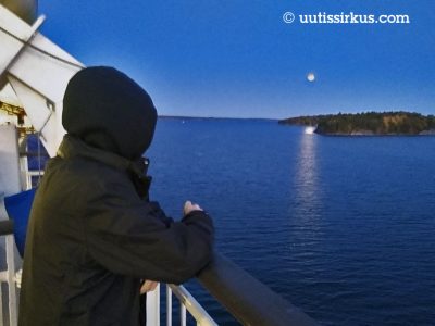 mustaan tuulitakkiin pukeutunut mies nojailee laivan reelinkiin huppu päässään hämärtyvässä illassa, kuu loistaa taivaalla miltei täytenä, meri on lähes tyyni