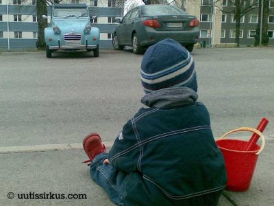 lapsi istuu jalkakäytävällä hiekkaämpäri ja lapio viesessään, katselee tien yli autoja, kadun toisella puolella puysäköitynä mm. Rättisitikka